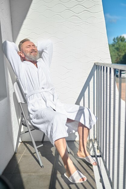 Entspannter Mann im weißen Bademantel und Pantoffeln auf dem Stuhl auf dem Balkon sitzend