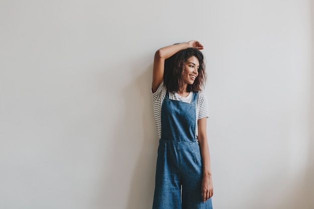 Entspanntes schwarzes Mädchen im Vintage-Denim-Gesamtaufstellung mit der Hand oben neben weißer Wand