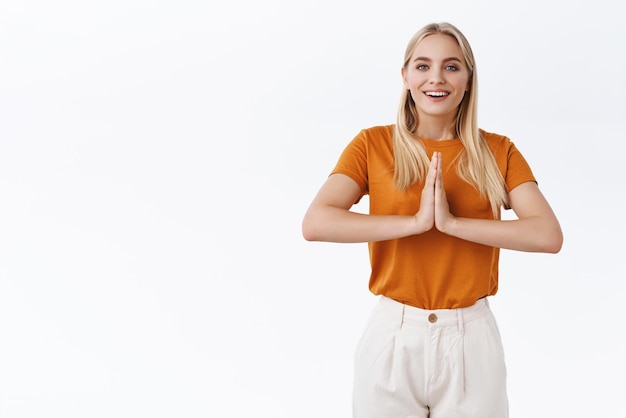Kostenloses Foto entspanntes und erleichtertes, gutaussehendes, modernes, blondes mädchen in orangefarbenem t-shirt hält die hände in gebetshandflächen, die in der nähe der brust zusammengepresst werden, als es sagt, dass namaste die yoga-atemstunde beendet und glücklich lächelt