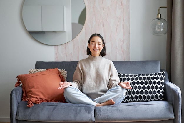 Entspannung und Geduld lächelnde junge Asiatin im gemütlichen Zimmer, die auf dem Sofa sitzt und meditiert