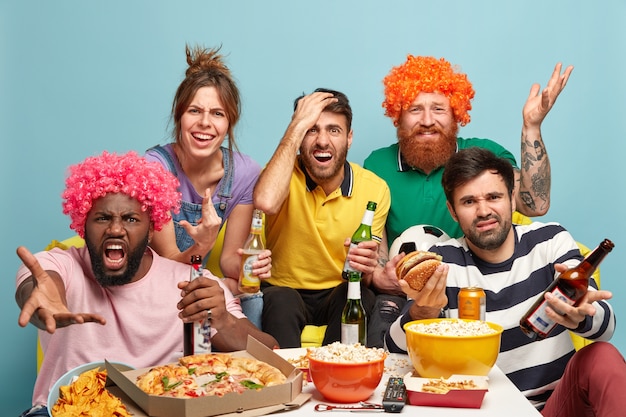 Enttäuscht schauen sich vier Männer und eine Frau ein Sportspiel an, sind unzufrieden mit Teamversagen, trinken Bier, essen einen Snack, drücken negative Reaktionen aus, haben schlechte Gefühle und posieren zu Hause zusammen auf dem Sofa. Team verlieren.