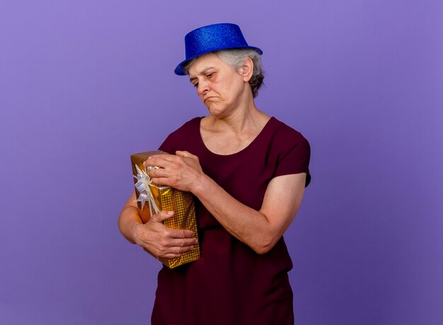 Enttäuschte ältere Frau, die Partyhut trägt, hält Geschenkbox lokalisiert auf lila Wand mit Kopienraum