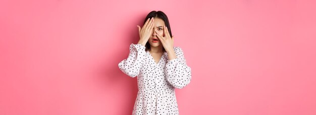 Enttäuschte asiatische Frau, die durch offene Augen der Finger späht und unzufrieden die Stirn runzelt und mit Disd anstarrt