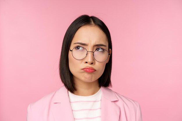 Enttäuschte asiatische Geschäftsfrau, Bürodame mit Brille, die verärgert auf etwas unfaires Schmollen schaut, das über rosa Hintergrund steht