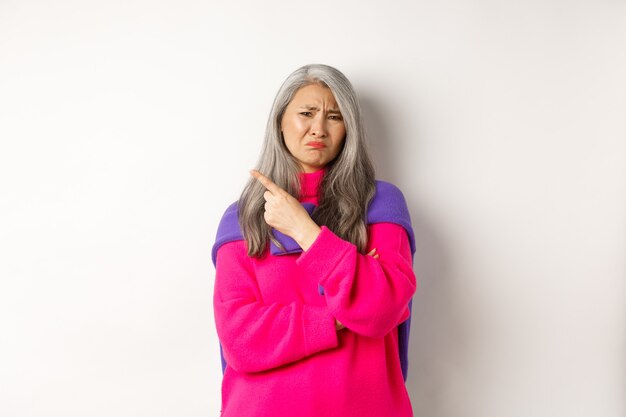 Enttäuschte asiatische Großmutter, die angewidert das Gesicht verzieht, mit dem Finger nach links zeigt und sich über etwas Schlechtes beschwert, steht auf weißem Hintergrund