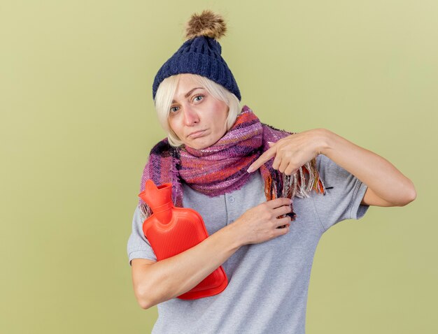 Enttäuschte junge blonde kranke slawische Frau, die Wintermütze und Schal hält und zeigt auf Wärmflasche lokalisiert auf olivgrüner Wand mit Kopienraum