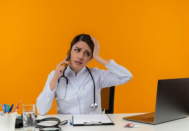 Enttäuschte junge Ärztin, die medizinische Robe und Stethoskop trägt, sitzt am Schreibtisch mit medizinischen Werkzeugen und Laptop, die am Telefon sprechen, das Seite lokalisiert auf gelber Wand betrachtet