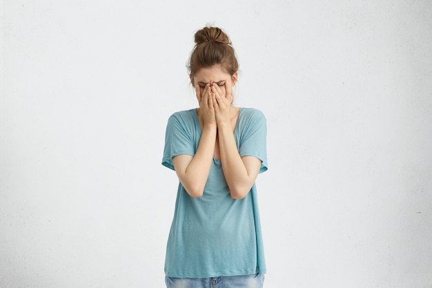 Enttäuschte, niedergeschlagene Frau mit Haarknoten, die ein blaues, lockeres T-Shirt trägt, das ihr Gesicht mit müden und erschöpften Händen bedeckt. Verzweifelte Frau, die Depression hat, die ihr weinendes Gesicht mit den Händen versteckt