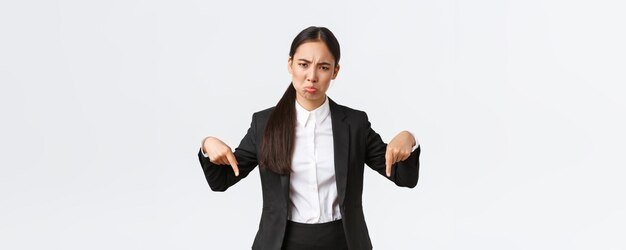 Enttäuschte, schmollende asiatische Geschäftsfrau, die sich über unfaire Situation beschwert Unternehmerin im Anzug, die mit den Fingern nach unten zeigt und das Gesicht verzieht, unzufrieden mit verärgertem weißem Hintergrund