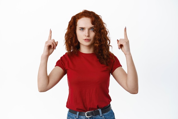 Kostenloses Foto enttäuschte, stirnrunzelnde frau braucht eine erklärung, die mit dem finger nach oben zeigt und ernsthaft in die kamera starrt und sich auf dem obersten banner beschwert, das vor weißem hintergrund steht