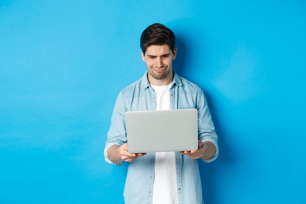 Enttäuschter, gutaussehender Mann, der auf den Laptop-Bildschirm schaut und das Gesicht verzieht, etwas Schlechtes im Internet beurteilt und auf blauem Hintergrund steht.