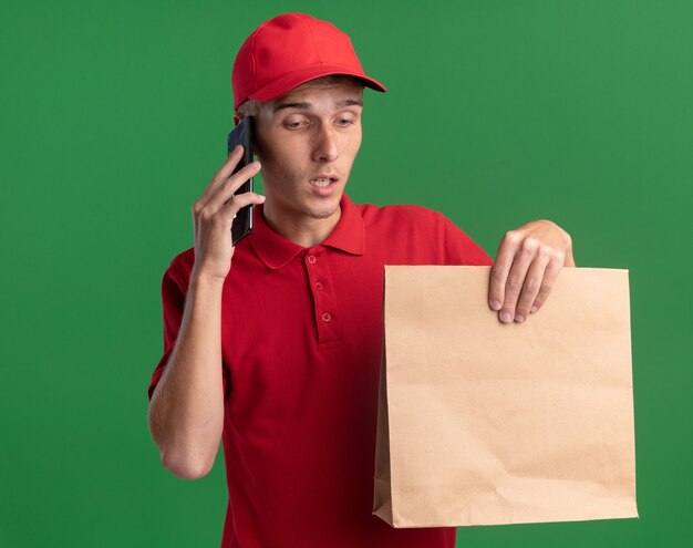 Enttäuschter junger blonder Lieferjunge hält Papierpaket und telefoniert