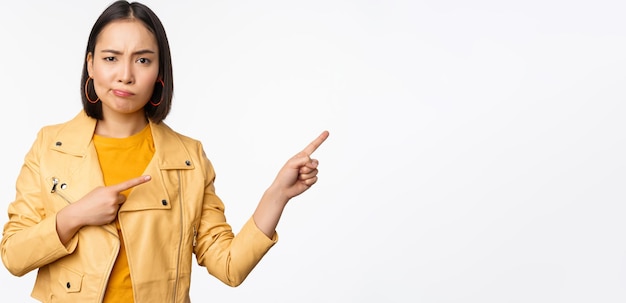 Enttäuschtes asiatisches Mädchen, das die Stirn runzelt und schmollend verärgert zeigt und mit dem Finger direkt auf die Werbung zeigt, die über weißem Hintergrund steht
