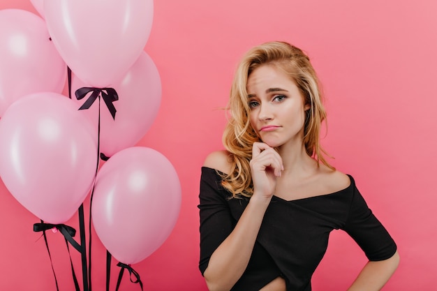 Enttäuschtes blondes Mädchen mit blauen Augen, die in der Nähe von Heliumballons stehen. Porträt der traurigen kaukasischen Dame lokalisiert auf rosa Wand während des Geburtstags-Fotoshootings.