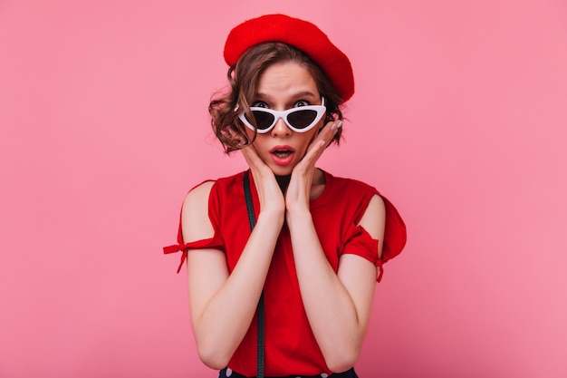Enttäuschtes französisches weibliches Modell, das in der Sonnenbrille aufwirft. unglückliche kurzhaarige Frau in der roten Baskenmütze isoliert.