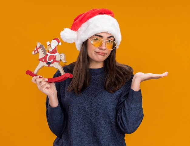 Enttäuschtes junges kaukasisches Mädchen in Sonnenbrille mit Weihnachtsmütze hält Weihnachtsmann auf Schaukelpferdedekoration und hält Hand offen auf orange Hintergrund mit Kopienraum