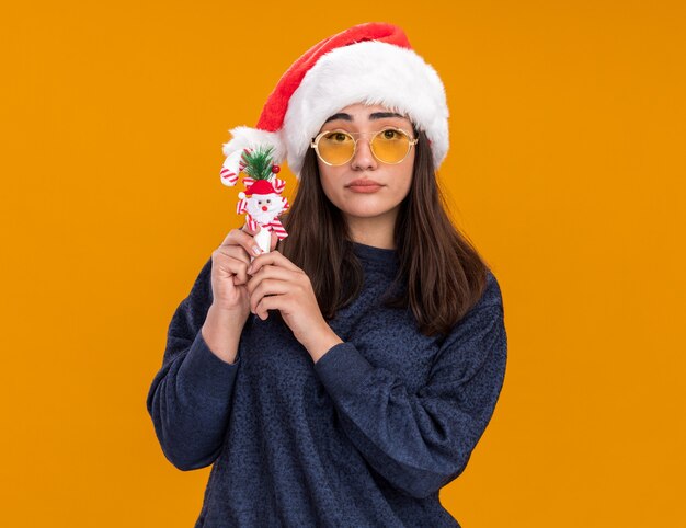 Enttäuschtes junges kaukasisches Mädchen in Sonnenbrille mit Weihnachtsmütze hält Zuckerstange isoliert auf oranger Wand mit Kopierraum