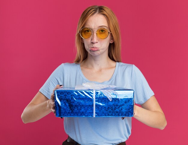 Enttäuschtes junges rothaariges Ingwermädchen mit Sommersprossen in Sonnenbrille mit Geschenkbox