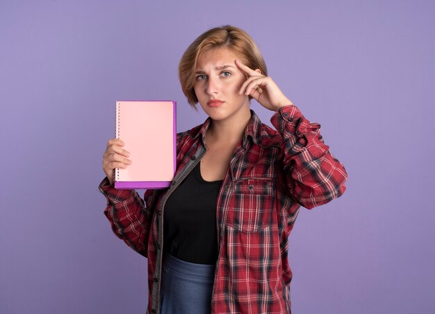 Enttäuschtes junges slawisches Studentenmädchen legt Hand auf Tempel hält Buch und Notizbuch