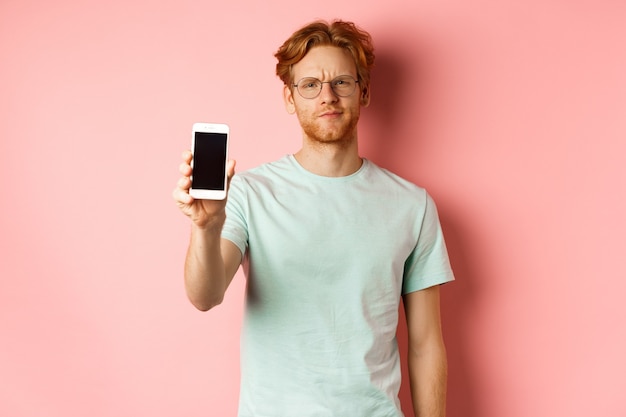 Enttäuschtes männliches Model runzelt die Stirn, zeigt Smartphone-Bildschirm und steht über rosa Hintergrund