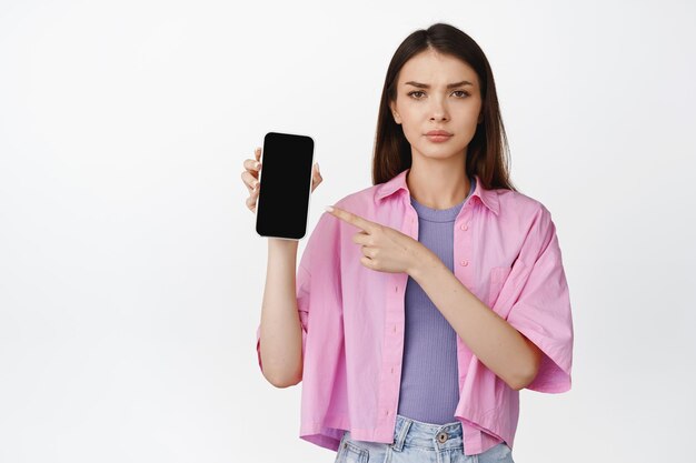 Enttäuschtes Teenager-Mädchen, das auf den Smartphone-Bildschirm zeigt und die Stirn runzelt, als Ausdruck von Abneigung gegen unzufriedene Emotionen, die über weißem Hintergrund stehen