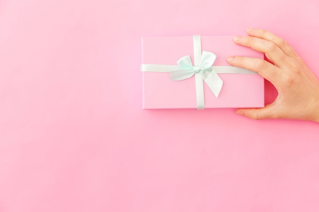 Entwerfen sie einfach die weibliche hand der frau, die rosafarbene geschenkbox einzeln auf rosafarbenem buntem modischem hintergrund hält. weihnachten neujahr geburtstag valentinstag feier präsentieren romantisches konzept. platz kopieren