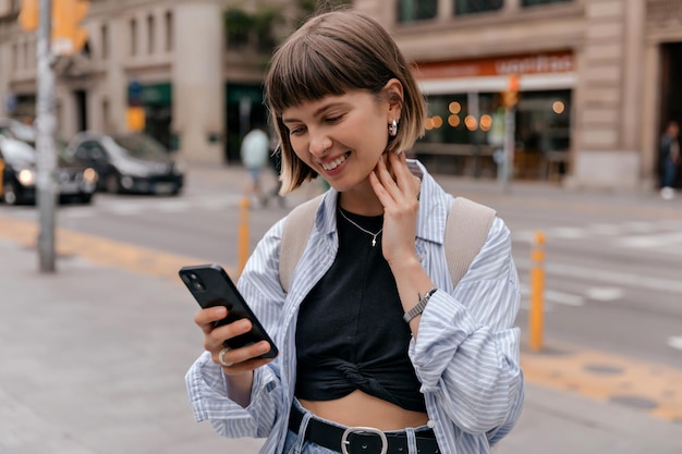 Entzückend lächelndes gerades kurzes Haar, das lächelt, während es das Smartphone in der Stadt hält und ein blaues Hemd und ein schwarzes Oberteil trägt Außenfoto einer kaukasischen Frau, die an warmen Tagen mit Rucksack in der Stadt spaziert