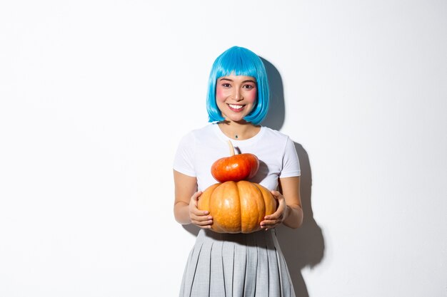 Entzückende asiatische Frau in der blauen Perücke, die zwei niedliche Kürbisse hält und in die Kamera lächelt und Schulmädchen-Outfit für Halloween-Party trägt.