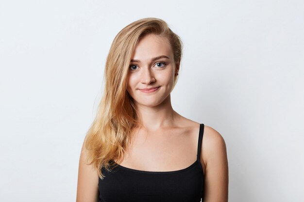 Entzückende blauäugige Frau mit langen glatten hellen Haaren, trägt lässiges schwarzes T-Shirt, schaut glücklich in die Kamera, als würde sie mit ihrem Freund spazieren gehen, hat gute Laune, isoliert auf weißem Studio