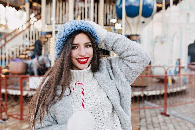 Entzückende dunkelhaarige Frau, die Freizeit im Vergnügungspark am Winterwochenende verbringt. Foto im Freien der atemberaubenden brünetten Dame im blauen Hut, der Weihnachtsbonbons nahe Karussell isst.