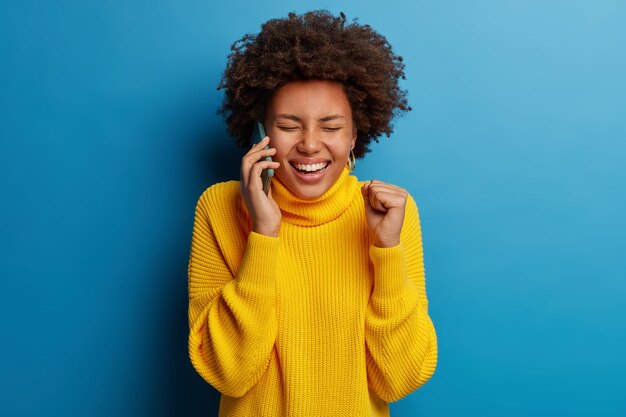 Entzückende dunkelhäutige erwachsene Frau gekleidet in gelbem Pullover unter Verwendung des Mobiltelefons mit einem glücklichen Ausdruck