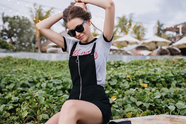 Entzückende europäische frau im weinlese-outfit, das mit ihrem dunklen haar auf natur spielt. foto im freien des fröhlichen weißen mädchens in der schwarzen sonnenbrille, die neben grünem rasen sitzt.