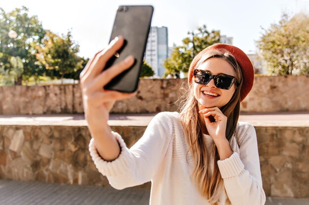 Entzückende europäische Frau in schwarzer Sonnenbrille, die an einem guten Herbsttag chillt Außenaufnahme einer glamourösen französischen Grl, die Selfie auf der Straße macht