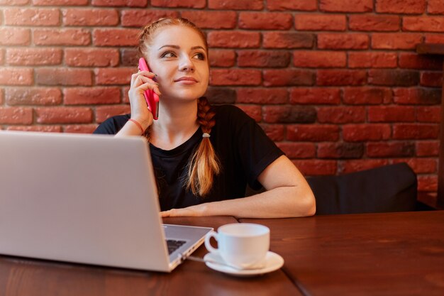 Entzückende Frau, die auf Smartphone im Café spricht, während sie online über Laptop arbeitet