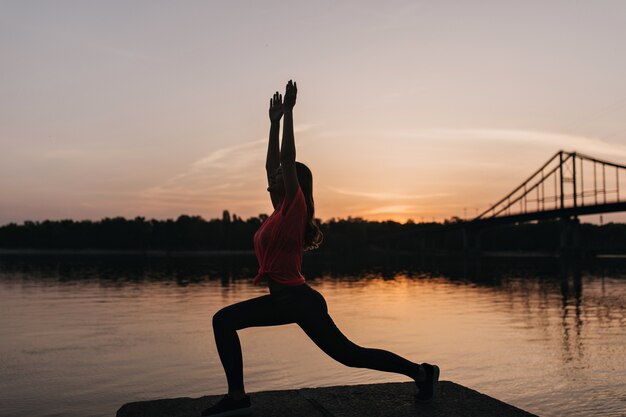 Entzückende Frau im rosa T-Shirt, das Yoga tut und Abendhimmel betrachtet. Außenporträt des charmanten weiblichen Modells, das Training nahe Fluss genießt.