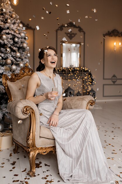 Entzückende Frau im silbernen Kleid sitzt vor einem Weihnachtsbaum mit einem Champagnerglas in ihrer Hand
