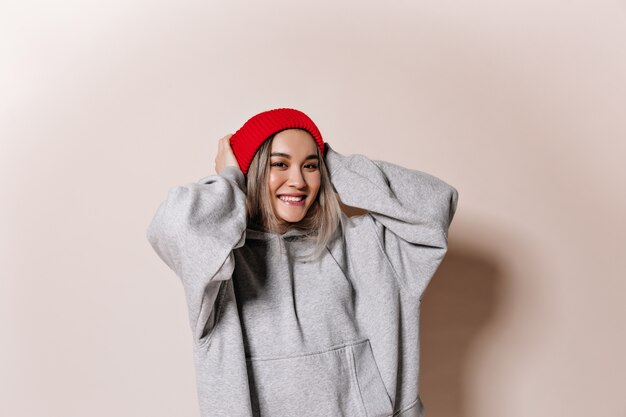 Entzückende Frau in der roten Kappe und im Sweatshirt, die auf isolierter Wand lächeln