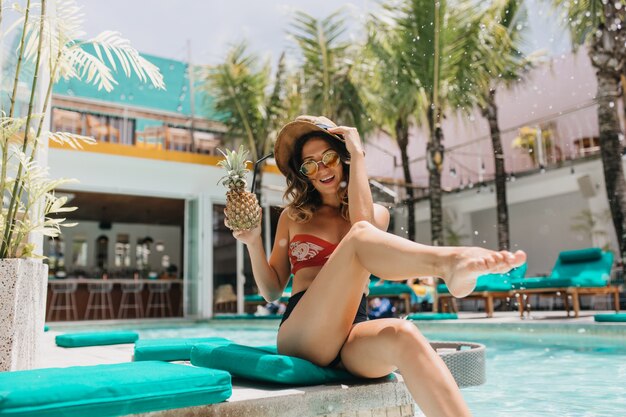 Entzückende Frau in der Sonnenbrille, die spielerisch am Pool aufwirft. Außenaufnahme der attraktiven lockigen Frau im Hut, der Ananas-Cocktail am exotischen Resort trinkt.