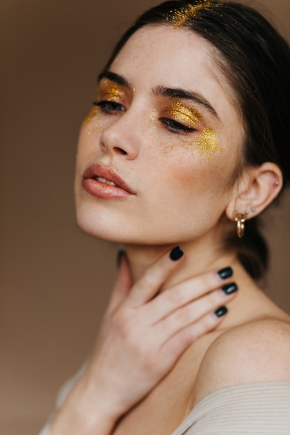 Entzückende Frau mit den schwarzen Haaren, die auf brauner Wand aufwerfen. Erstaunliches weibliches Modell mit goldenem Make-up, das mit geschlossenen Augen steht.