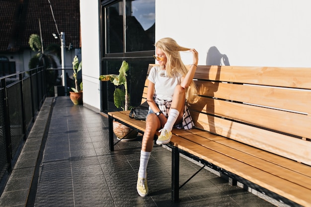 Entzückende gebräunte Frau in den weißen Socken, die mit langen blonden Haaren spielen. Außenporträt des glückseligen kaukasischen Mädchens in den gelben Schuhen, die auf hölzerner Bank abkühlen.