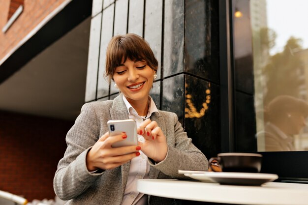 Entzückende junge brünette Dame in der grauen Jacke und im weißen Hemd unter Verwendung des Smartphones und arbeitet im Laptop