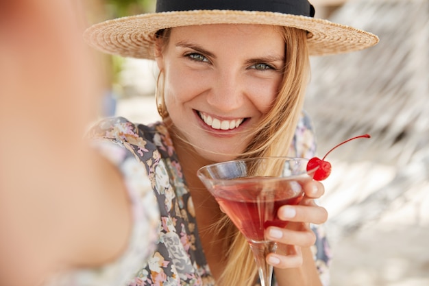 Entzückende junge Frau mit glücklichem Ausdruck, hält frischen Kirschcocktail, macht Selfie, posiert im Freien, hat entzückenden Blick, froh, Sommerruhe im Ferienland zu haben. Glücks- und Erholungskonzept