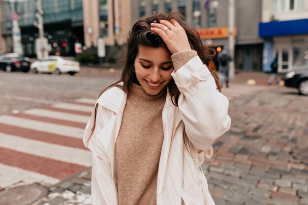 Entzückende junge stilvolle Frau, die in modischem Mantel auf der Straße steht. Sie hat ein sanftes Lächeln und berührt ihr Haar, während sie nach unten schaut