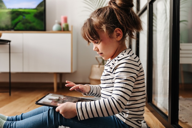 Entzückende kleine Frau in guter Laune sieht Cartoons in Tablette und Posen im Wohnzimmer.