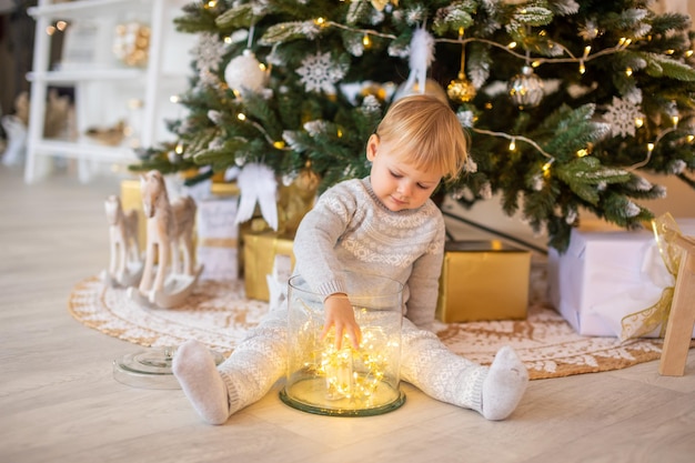 Entzückendes baby, das in der nähe des weihnachtsbaums mit festlichen lichtern und weihnachtsgeschenken weihnachten und neujahr sitzt