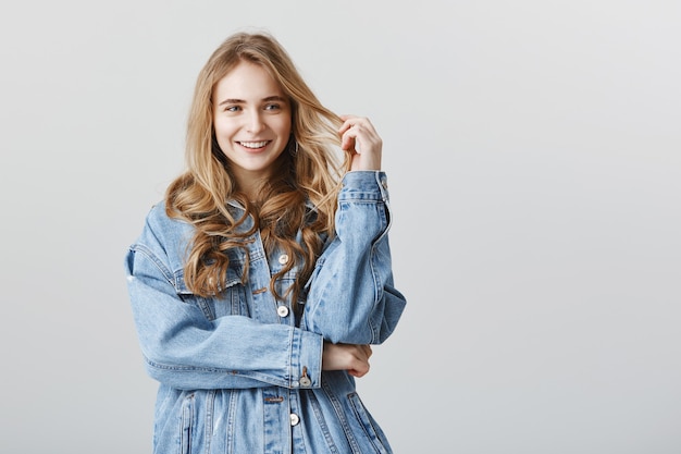 Entzückendes blondes Mädchen, das glücklich in der Jeansjacke lächelt und über graue Wand steht