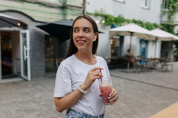 Entzückendes hübsches Mädchen in weißem T-Shirt mit Smoothie schaut weg und lächelt auf der hellen Sommerstraße in der Stadt Glückliches Mädchen geht an guten Tagen auf der Straße