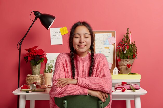 Entzückendes junges Mädchen mit asiatischem Aussehen, zwei Zöpfen, hält die Augen geschlossen, während es auf dem Stuhl sitzt, stellt sich vor, dass während der Weihnachtszeit etwas Wunderbares passiert ist