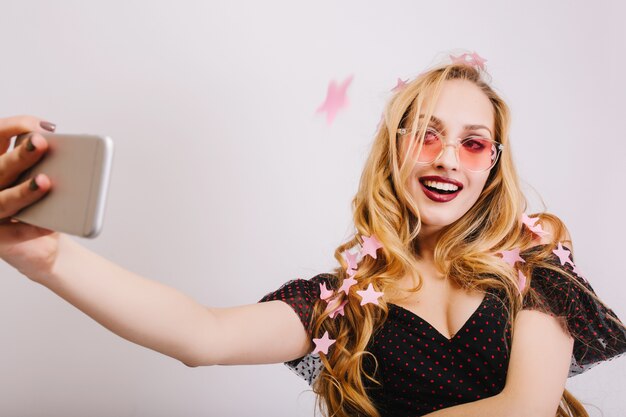 Entzückendes junges Mädchen mit dem blonden langen lockigen Haar, das Selfie auf der Partei nimmt, lächelnd, bedeckt mit rosa Sternenkonfetti. Trage bunte Brille, schwarzes Kleid.