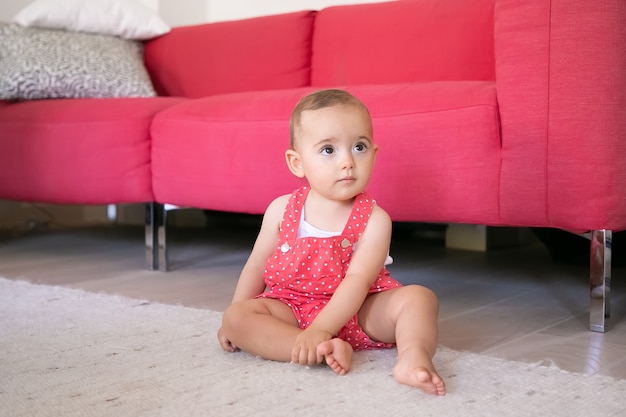 Entzückendes kleines Baby, das auf Teppich barfuß im Wohnzimmer sitzt. Lustiges nachdenkliches Mädchen in roten Latzhose, die jemanden ansieht und ihr Bein berührt. Wochenende, Kindheit und Zuhause sein Konzept